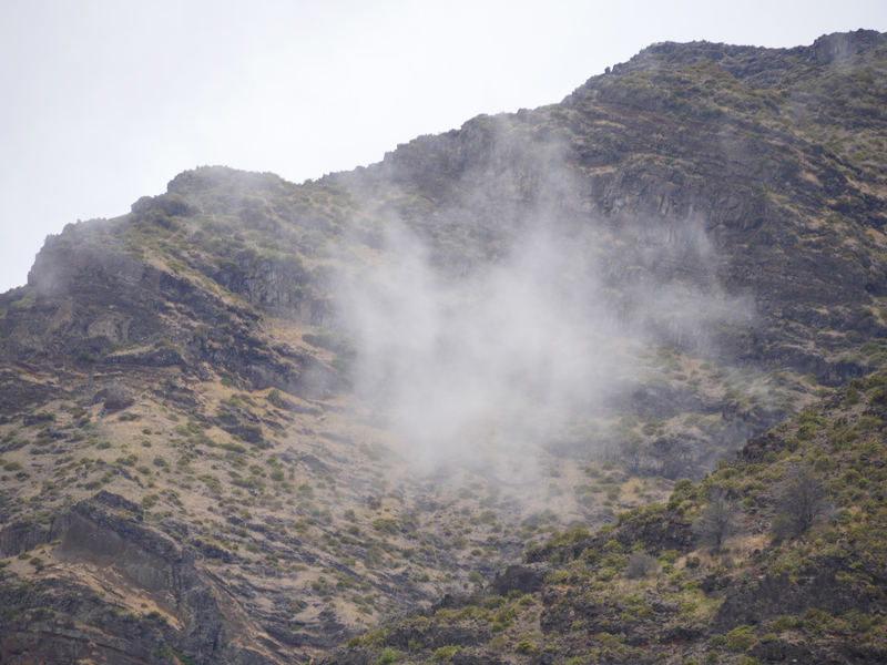 A cloud hangs in the air, backed by a steep rocky hill in Hawaii