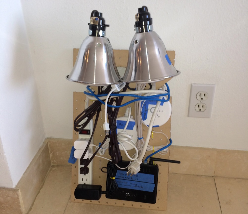 A pegboard test rig of consumer networking hardware and two lightbulbs sits on a tile floor beside an outlet